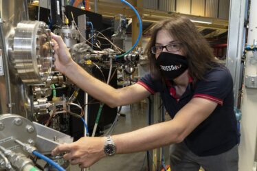 Photo of Jonathan Pelliciari in a lab, wearing a face mask and holding onto a large instrument with many metal knobs and wires.