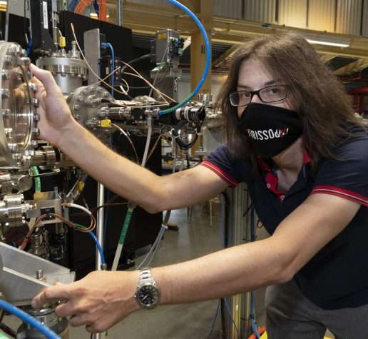 Photo of Jonathan Pelliciari in a lab, wearing a face mask and holding onto a large instrument with many metal knobs and wires.