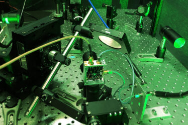 Photo of a tabletop experimental setup featuring lasers, mirrors, and other small, black or metal parts that are screwed into a large metal plate covered with screw openings. The entire scene is tinted green due to the green laser light being used.