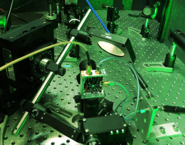 Photo of a tabletop experimental setup featuring lasers, mirrors, and other small, black or metal parts that are screwed into a large metal plate covered with screw openings. The entire scene is tinted green due to the green laser light being used.