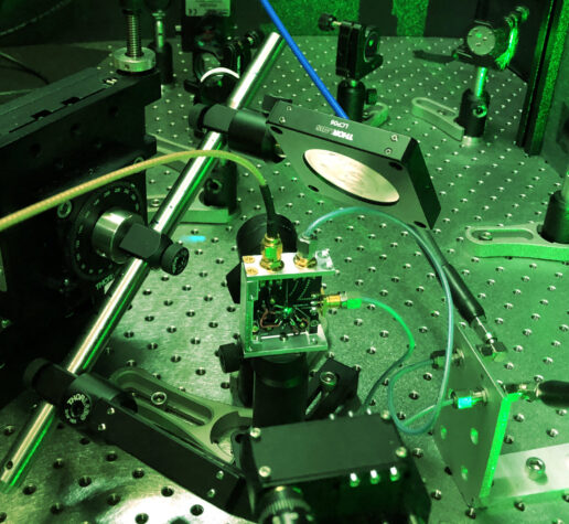 Photo of a tabletop experimental setup featuring lasers, mirrors, and other small, black or metal parts that are screwed into a large metal plate covered with screw openings. The entire scene is tinted green due to the green laser light being used.