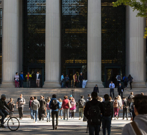 Photo of students crossing the street at 77 Mass. Ave., in front of MIT's main entrance, which features steps, large columns, and glass windows.