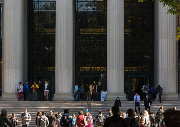 Photo of students crossing the street at 77 Mass. Ave., in front of MIT's main entrance, which features steps, large columns, and glass windows.