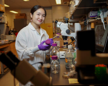 Catherine Ji, in the lab with purple gloves and surrounded by microscopes and lab equipment, labels a glass bottle.