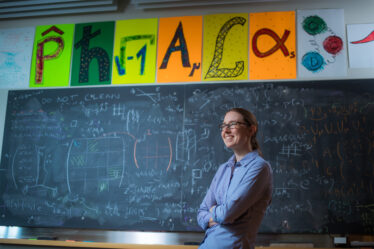 Professor Phiala Shanahan posing in front of chalkboard filled with equations and her name spelled out with scientific symbols.