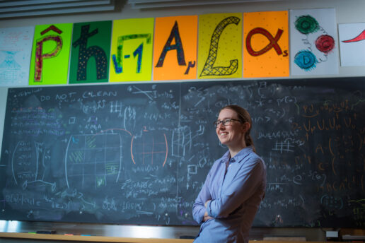 Professor Phiala Shanahan posing in front of chalkboard filled with equations and her name spelled out with scientific symbols.