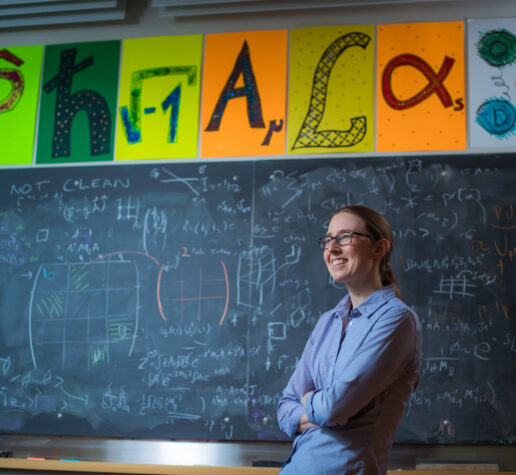 Professor Phiala Shanahan posing in front of chalkboard filled with equations and her name spelled out with scientific symbols.