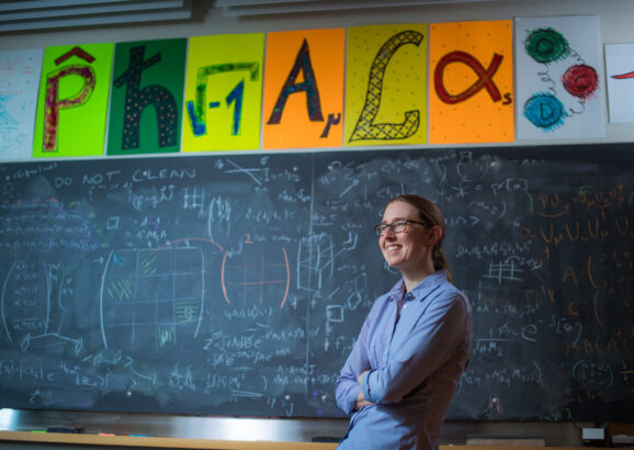 Professor Phiala Shanahan posing in front of chalkboard filled with equations and her name spelled out with scientific symbols.