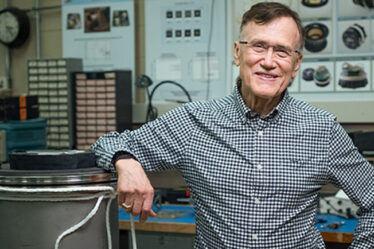 George Ricker stands in a lab, resting his elbow on a large metal cylinder