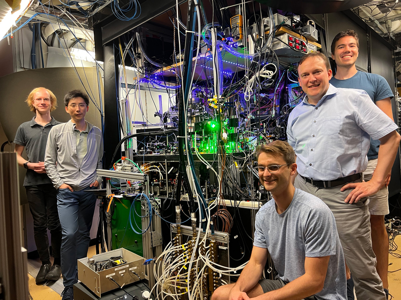 The 5 people stand around a large, crowded machine, full of dangling wires, green lights, blue LEDS.