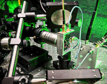 A closeup of the laser equipment shows metallic rods, a circuit board, wires, and lenses, all in a green glow.