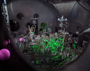 The inside of a LIGO chamber with an array of lenses, metallic pieces, and cords. There is green and pink lighting.