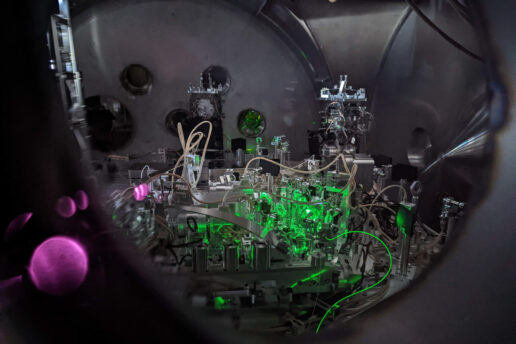 The inside of a LIGO chamber with an array of lenses, metallic pieces, and cords. There is green and pink lighting.