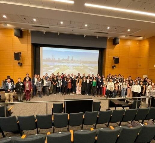 About 65 people pose standing in a semicircle on a large auditorium stage, with the backs of empty seats in the foreground and a video screen behind them.