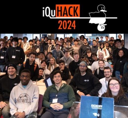 Perhaps 150 people pose for a photo in a large classroom. About a third are seated in the foreground and the rest stand. At top is a banner that reads iQuHACK 2024, with the image of two nearly identical ducks, one alive and one shown as dead with X's for eyes.
