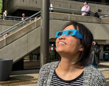 Joanna Chen wears special eclipse glasses and look up, with other people doing the same in background.