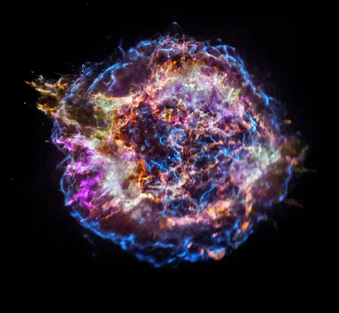 In the center of a black background is a large circular object outlined by electric blue filaments that jolt and weave throughout the structure. The object, which is the supernova remnant Cassiopeia A, includes strings of vibrant colors including purple, orange, yellow and blue.