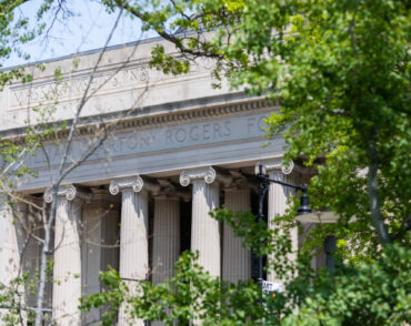 The columns of Building 7 are visible through green foliage on a sunny Spring day.