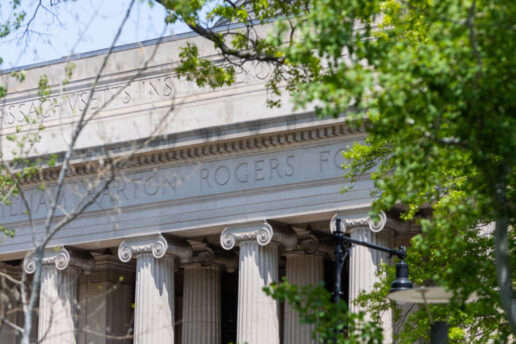 The columns of Building 7 are visible through green foliage on a sunny Spring day.