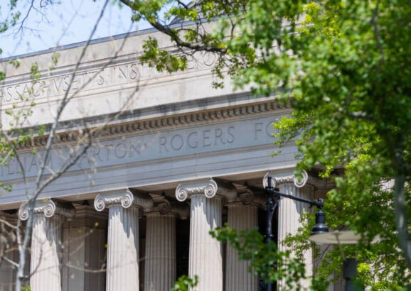 The columns of Building 7 are visible through green foliage on a sunny Spring day.