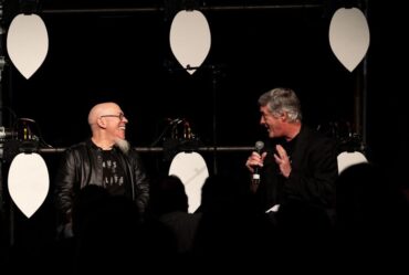 Jordan Rudess and Joseph Paradiso speak to each other on a black stage with white petals interspersed among them