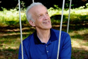 Daniel Kleppner, a white-haired man wearing a blue short-sleeved shirt, sits on what appears to be some kind of swing.