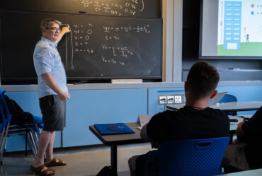 Professor stands at a chalkboard, lecturing in a classroom