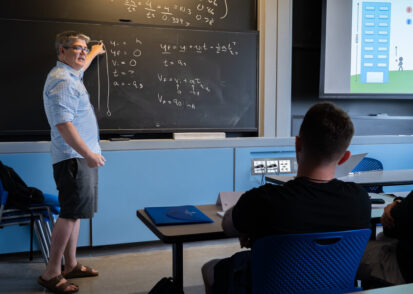 Professor stands at a chalkboard, lecturing in a classroom