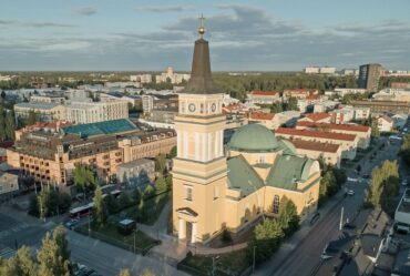 Aerial view of a yellow church with a green dome and tall steeple in the center of a town, surrounded by buildings and trees.