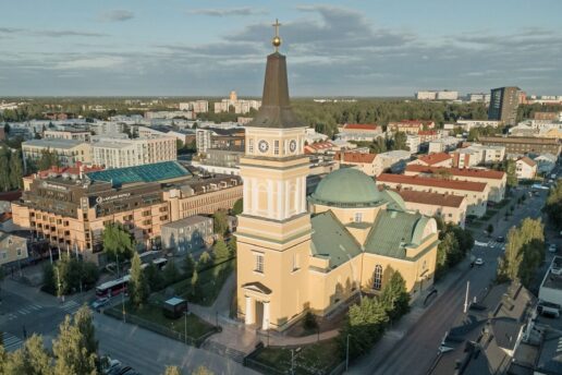 Aerial view of a yellow church with a green dome and tall steeple in the center of a town, surrounded by buildings and trees.