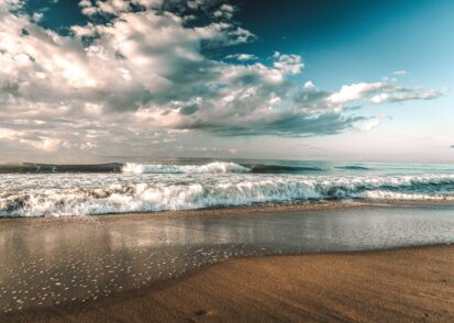 Gentle ocean waves breaking on a sandy shore under a cloudy blue sky.