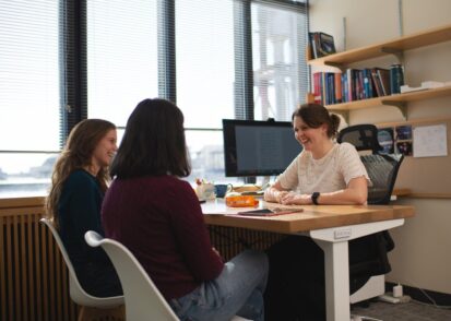 Anna-Christina Eilers sits at her desk, smiling warmly at two female students who sit facing her