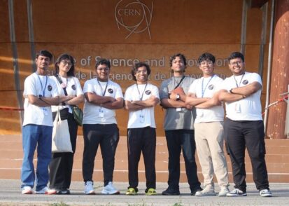 Manu Srivastava and 6 high school students pose standing in front of a sign at CERN with their arms folded