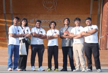 Manu Srivastava and 6 high school students pose standing in front of a sign at CERN with their arms folded