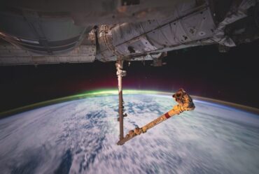 Panoramic orbital view of Earth's curved horizon illuminated by a green aurora layer, with the Canadarm2 robotic arm extended from the International Space Station in the foreground.