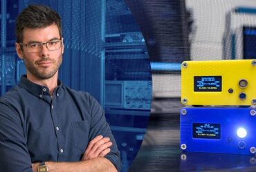 Man posing with arms crossed looking straight at camera stands on left of image. On right is image of two rectangular particle detectors one on top of the other. The top one is yellow, the bottom is blue. The blue one has a light that is lit.