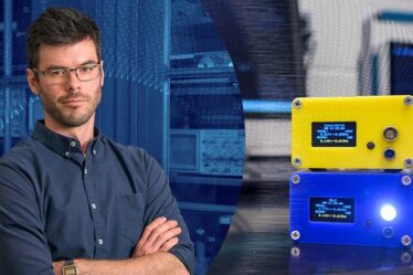 Man posing with arms crossed looking straight at camera stands on left of image. On right is image of two rectangular particle detectors one on top of the other. The top one is yellow, the bottom is blue. The blue one has a light that is lit.