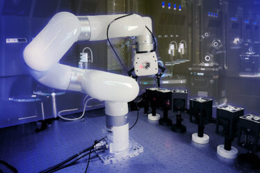 A white robotic arm lines up black boxes in an experiment. A rendering of a futuristic robot lab is in the background.