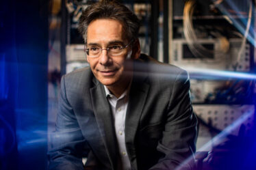 Portrait of MIT Professor William D. Oliver wearing glasses and a grey suit jacket, looking to the side in front of rack-mounted laboratory equipment with blue lighting effects.