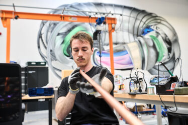 Francesco Sciortino, wearing gloves, looks down a long copper-colored rod. Various devices can be seen on workbenches behind them and there is a large mural on the wall in the background that appears to represent a stellerator.