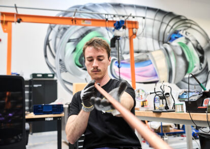 Francesco Sciortino, wearing gloves, looks down a long copper-colored rod. Various devices can be seen on workbenches behind them and there is a large mural on the wall in the background that appears to represent a stellerator.
