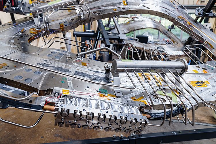 A close-up view of intricate metallic modular sections, piping, and alignment mechanisms of a stellarator fusion device under assembly in a workshop.